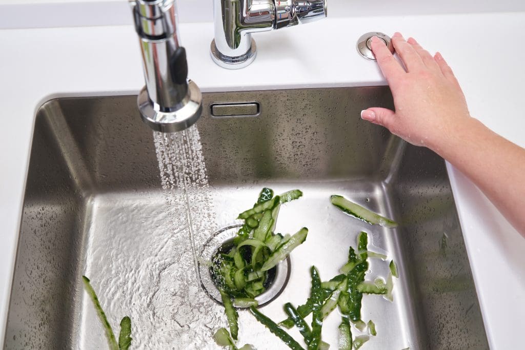 A drain with green food scraps and a person pressing a button for the garbage disposal
