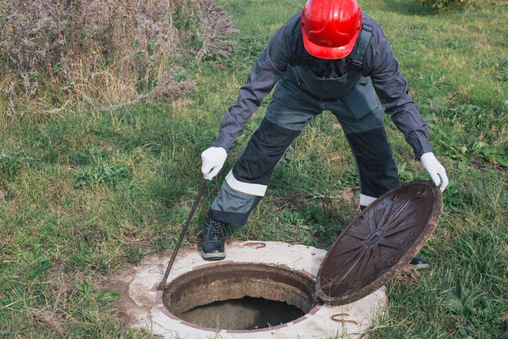 Worker in safety gear opening septic tank lid with metal rod during inspection on grassy field.