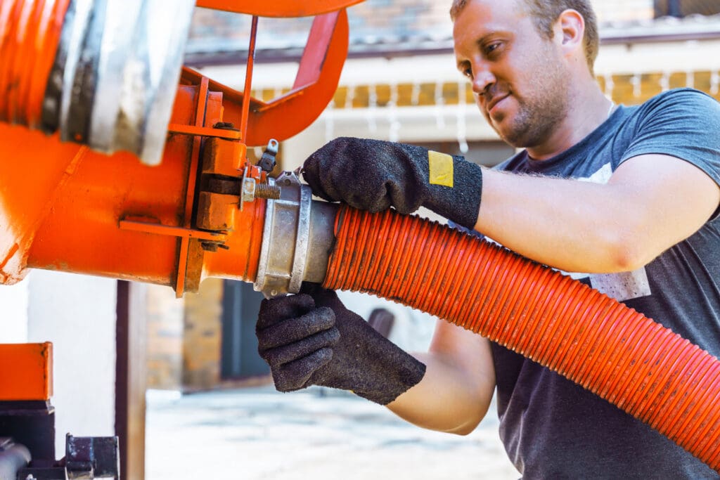 A worker connecting a large orange industrial hose to equipment while wearing safety gloves.