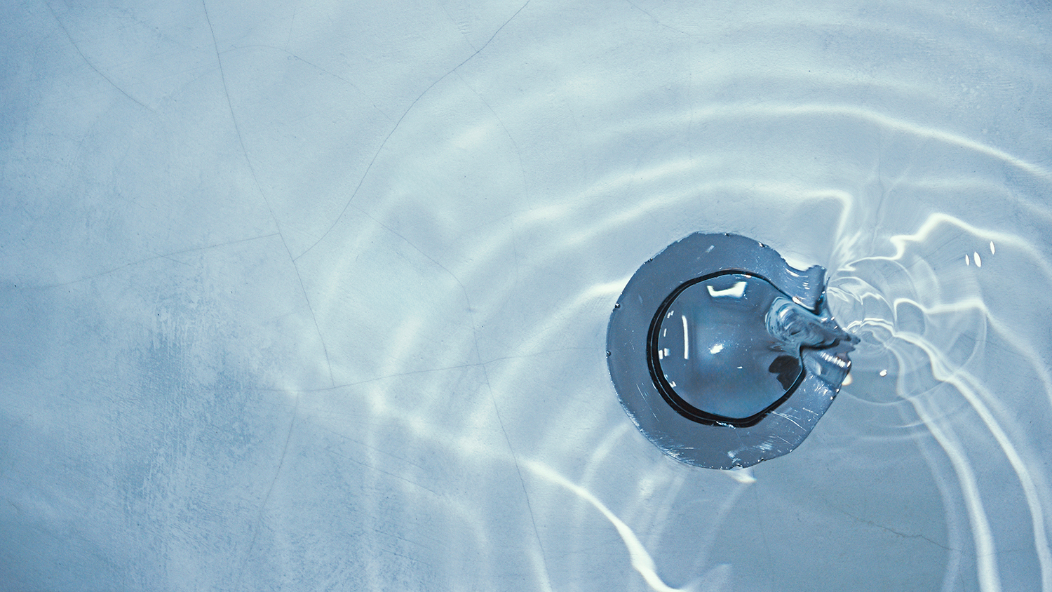 A bathtub drain, with blue tinged water rippling over it.